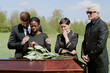 © Seventyfour - Black woman crying while holding lilies, comforted by Caucasian man, standing beside middle aged Caucasian woman and senior Caucasian man near coffin during funeral outdoors
