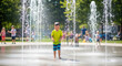 © Shootdiem - Smiling young boy enjoying a sunny day playing among water fountains at a splash pad, surrounded by blurred kids and greenery.