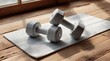 © asAI12 - Two light-grey dumbbells rest on a textured white mat atop a rustic wooden floor, bathed in sunlight streaming from a nearby window