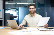 © Tetiana - Portrait of a smiling young man working in the office with documents, typing on a laptop and looking confidently at the camera