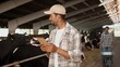 © VAKSMANV - Joyful African American man farmer in cap typing on tablet standing in cowhouse. People working on background. Cattle in stall. Male using gadget in farming in barn. Cows in stable. Dairy business