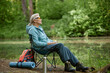 © Seventyfour - Senior Caucasian woman sitting on folding chair near forest lake holding cup, wearing sunglasses, relaxing outdoors with backpack and yoga mat beside, enjoying peaceful nature surroundings