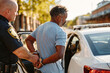 © Alina - A police officer places handcuffs on a man outside a parked police vehicle in a city setting.