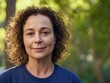 © tao - Middle-aged woman with curly brown hair smiles at the camera wearing a blue T-shirt standing in front of a green tree with sunlight shining on her face, capturing a warm and natural outdoor moment.