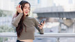 © Exnoi - Young Asian woman enjoying music with headphones and smartphone in a modern city. Relaxed and stylish, she represents digital lifestyle, tech-savvy youth, and urban audio streaming culture.