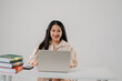 © pichet - Young woman smiling while working on laptop at desk with books and smartphone in minimalist office setting presenting a cheerful, productive atmosphere