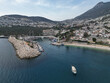 © MikeDrone - Aerial view of Kas marina in Antalya, Turkiye, boats along the quay beside a rock breakwater and white lighthouse, teal Mediterranean water, hillside town on green slopes, warm late afternoon light