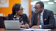 © Samady - Two professionals engaged in a discussion at a desk with a laptop, wearing formal attire and glasses, in a modern office setting.