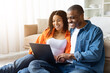 © Anastasiya - Smiling young African American couple sitting on floor with laptop at home, browsing together and enjoying time in casual relaxed setting, copy space