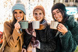 © Lomb - Three diverse friends looking at camera with mugs at a Christmas market. Happy multicultural group toasting with hot chocolate and enjoying the festive holiday season at night.