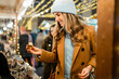 © Lomb - Woman shopping at a Christmas market, choosing a small golden gift box. Happy young customer buying presents for the holiday season at a festive outdoor fair at night.