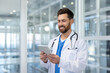 © Liubomir - A healthcare professional wearing a white coat and stethoscope smiles, using a digital tablet in a contemporary medical environment, showcasing technology integration in modern medicine.