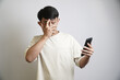 © yashis007 - A shocked Asian man in a beige t-shirt covers his face with his hand while looking at his smartphone, reacting to something surprising or embarrassing.