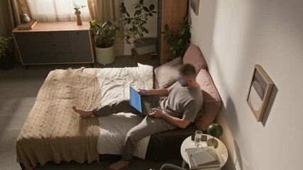 High-angle time lapse shot of young Caucasian man lying and sitting on bed at home, working on laptop computer, talking on phone in daytime, reading book in evening, turning off light, going to sleep