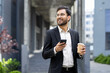 © Liubomir - A professional man in glasses and a suit stands outdoors holding a coffee and smartphone, smiling confidently. The background features a sleek, modern architectural design and blurred greenery.