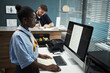 © DragonImages - Young Black woman working at computer in bank office, standing at desk and typing on keyboard while middle aged Caucasian man writing in background, modern workplace setting