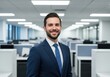 © PP Creative - A smiling businessman in a suit and tie stands confidently in a modern office environment with rows of desks and computers