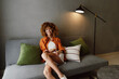 © SHOTPRIME STUDIO - Happy young woman with curly hair sitting on a gray sofa, wearing a stylish outfit, creating a cozy atmosphere in a modern interior