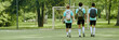 © Seventyfour - Header of three teenage boys, two Caucasian and one Black, walking together on outdoor soccer field carrying backpacks and soccer ball, heading toward goalpost with green trees in background