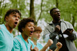 © Seventyfour - Group of multiethnic teenage boys cheering with excited expressions outdoors, Black man coach standing behind holding digital tablet and shouting encouragement during sports event
