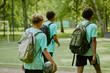 © Seventyfour - Three teenage boys, one Caucasian and two Black, walking together on outdoor soccer field carrying backpacks, one holding soccer ball, heading away from camera toward green park area