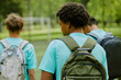 © Seventyfour - Group of teenage boys with backpacks walking outdoors on grassy field, multiethnic teenagers moving away from camera, casual clothing, blurred background with trees and soccer goal