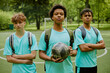 © Seventyfour - Three teenage boys standing on outdoor soccer field, two Caucasian and one Black, all wearing sports uniforms and backpacks, central boy holding soccer ball, looking confidently ahead
