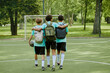 © Seventyfour - Three teenage boys, two Black and one Caucasian, walking together on outdoor soccer field with arms around each other, carrying backpacks, one holding soccer ball, viewed from behind