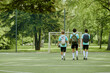 © Seventyfour - Three teenage boys walking together on outdoor soccer field carrying backpacks and soccer ball, heading toward goalpost surrounded by green trees