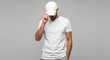 © Pindo - A man in a white t-shirt and cap adjusts his headwear, looking down against a neutral background.