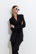© SHOTPRIME STUDIO - Stylish woman in black suit wearing white sunglasses stands confidently with arms crossed against white background, showcasing modern fashion and minimalistic elegance.