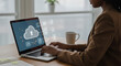 © Muhammad - Person using a laptop with cloud storage interface and a mug on the desk near a bright window