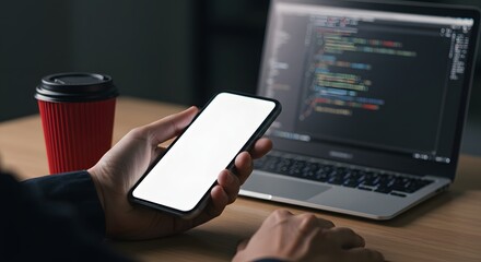 Developer using mobile phone with blank screen, laptop with code on desk