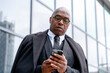 © Iryna - Business professional checks smartphone while standing outside a modern office building during the day