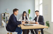 © Studio Romantic - Three colleagues in formal attire sit at a wooden table in a bright modern office, reviewing documents and laptops. Focused teamwork drives discussion and planning. Modern space supports meeting.