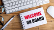 © Iryna - Welcome on board text on spiral notebook with pen, keyboard and mouse on wooden desk