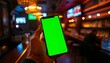 © Chaweewan - Hand holding smartphone with green screen in dimly lit bar, setting, leisure