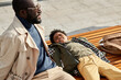 © TrueFrame Collective - Portrait of handsome African American father and son spending time together outdoors relaxing on bench enjoying sunny weather