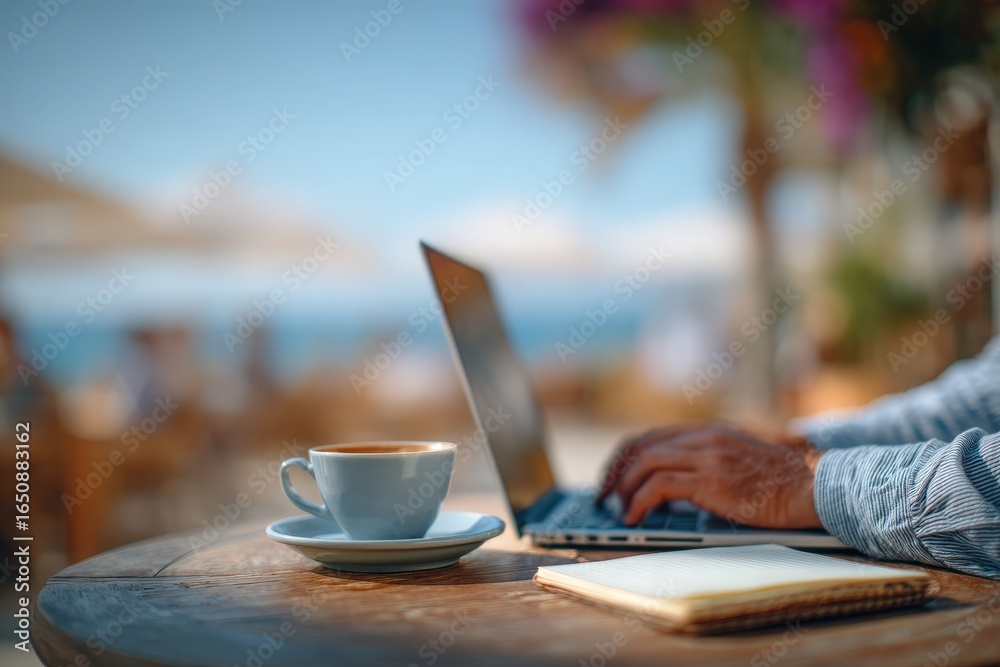 A person is typing on a laptop placed on a wooden table by the beach, enjoying coffee. The beach atmosphere has bright sunlight and blooming flowers in the background.