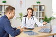 © Studio Romantic - Cheerful friendly female doctor at medical office in hospital showing examination results on laptop monitor screen and giving consultation to a young man patient sitting in clinic.