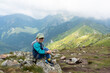 © Maryia - Happy young boy in cap and blue jacket sitting on rocky mountain summit with spectacular alpine valley panorama on cloudy day. Concept of family hiking, childhood adventure and mountain achievements.