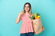 © luismolinero - Young woman holding a grocery shopping bag isolated on blue background giving a thumbs up gesture