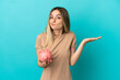 © luismolinero - Young woman holding a piggybank over isolated blue background having doubts while raising hands