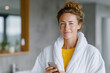 © AnaMadzar - Smiling woman wearing bathrobe holding smartphone in bathroom