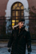 © Svetlana - Man in black winter coat with fur hood and gloves standing outdoors near brick building. Warm clothing, urban autumn fashion, casual walk, modern city style, architectural background