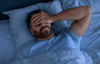 © Prostock-studio - Insomnia Problem. Man Covering Face With Hand Lying Unable To Sleep In Bed At Home, Suffering From Headache And Sleeplessness In Modern Bedroom Interior. Top View, Low Light