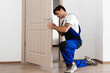 © Prostock-studio - Professional Repair Service Concept. Focused young male locksmith workman in blue uniform installing door knob. Installation of a lock on the front wooden entrance door at home or office workplace