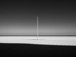 © Sergey - A minimalist black and white photo of a lone vertical pole on a vast, flat surface, casting a long shadow, with a stark horizon and shaded foreground.