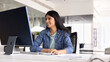 © fizkes - Positive young Indian professional woman in casual working on online project, Internet startup at computer monitor at workplace, sitting at office table in workspace alone, using mouse, smiling