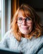 © Kowit - A smiling woman with red hair and glasses sits comfortably in a cozy setting, exuding warmth and approachability.
